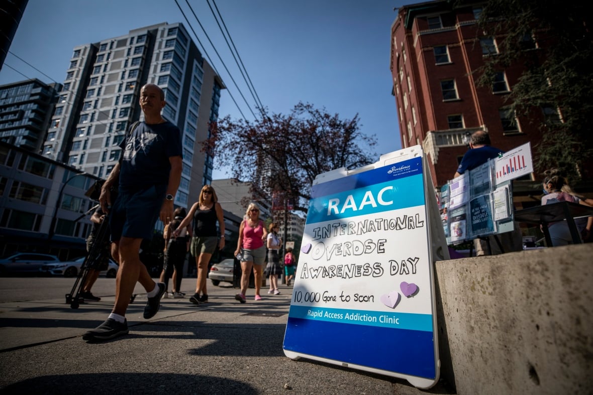 A person passes by a sandwich board that reads 'International Overdose Awareness Day, 10,000 gone too soon' on a sunny day.