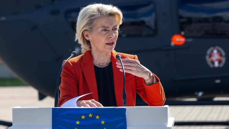 A woman stands at a podium, speaking into a microphone. She appears to be outside, with some sort of large vehicle behind her. The EU flag is on the podium. 