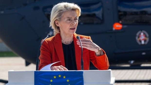 A woman stands at a podium, speaking into a microphone. She appears to be outside, with some sort of large vehicle behind her. The EU flag is on the podium. 