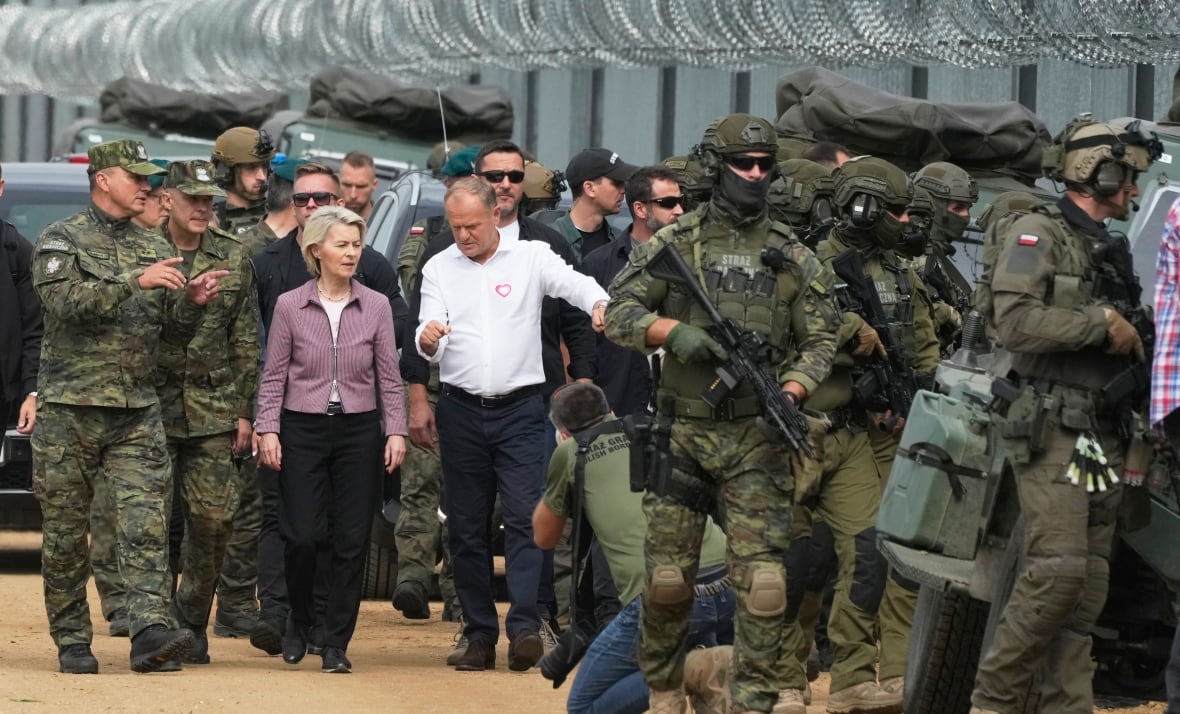 A woman and a man walk together alongside a fence topped with circular barbed wire. A number of soldiers surround them.