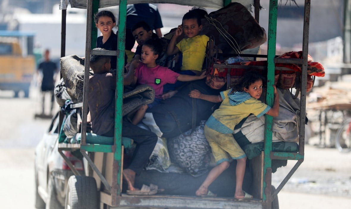 Children cling to the back of a vehicle loaded with blankets and luggage. 