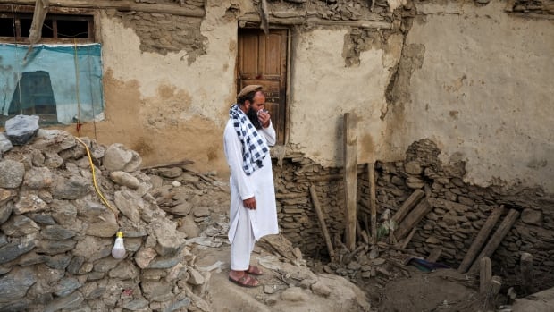 A man in white clothing stands looking bereft in the midst of the rubble of a collapsed building.