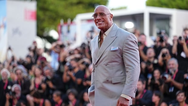 A bald man wearing glasses and a light grey suit smiles as he poses for a photograph, outside on a red carpet, with a large crowd of photographers behind him. 