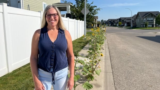A woman wearing glasses stands in front of a row of large sunflowers growing from a small crack of the sidewalk next to the road. 