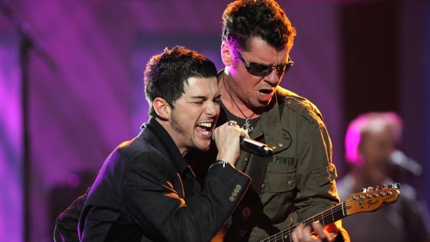A man wearing a black blazer sings into a microphone, while standing beside a man playing guitar who is wearing sunglasses.