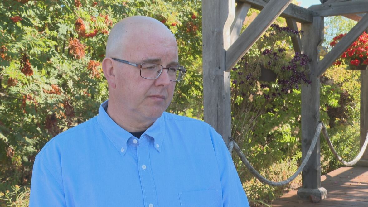 A man wearing a blue collared shirt looks off camera; there is greenery in the background