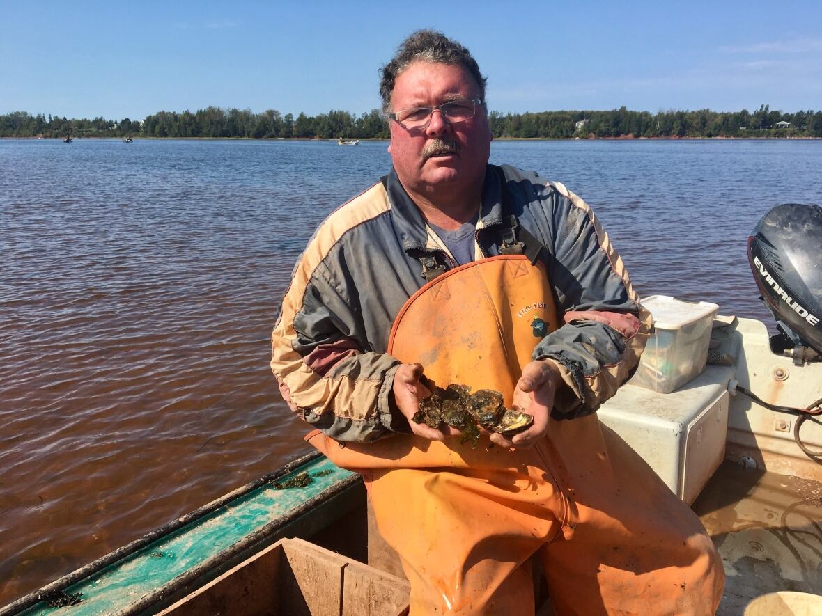 A man sits on a boat in the water, holding up wild oysters.