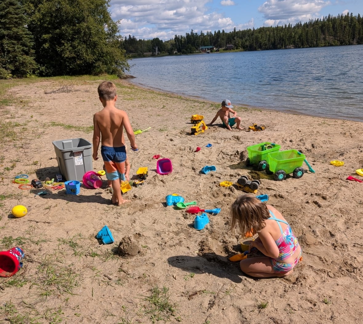 Three children play with shovels, buckets and other toys on the beach of a lake surrounded by trees on a sunny summer day.