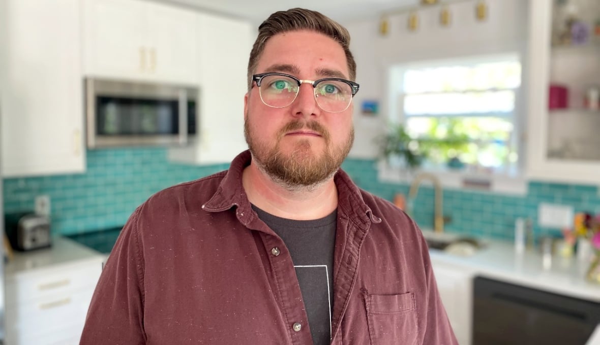 A white man with short blonde hair and a beard wear glasses and a black shirt under a red shirt with buttons. He is in a kitchen