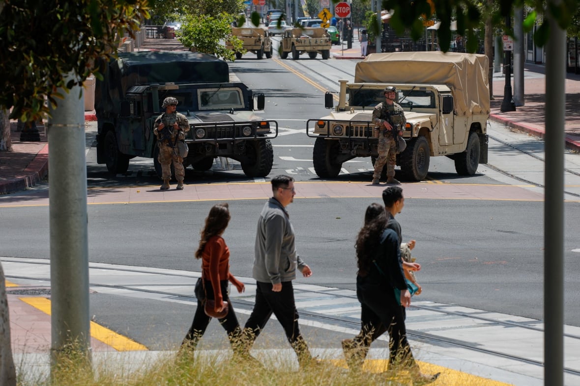 Civilians walk through a cross corridor while military figures look at Kamu with large weapons.