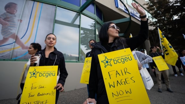 People walk with yellow sandwich boards reading 'Fair Deal Fair Conditions!! On Strike' and 'Fair Wages Now'.