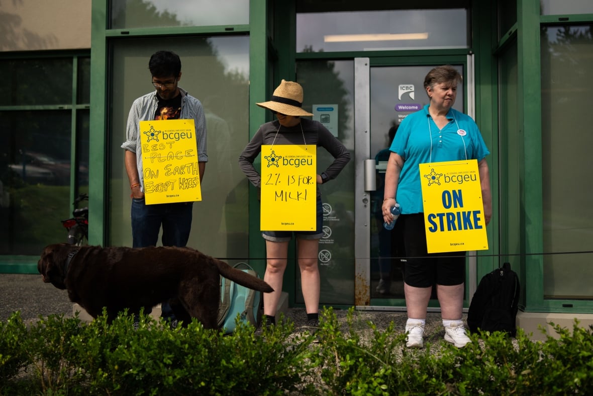 People stand outside an office with yellow sandwich boards reading 'Best place on earth except wages', '2% is for milk', and 'On Strike'.