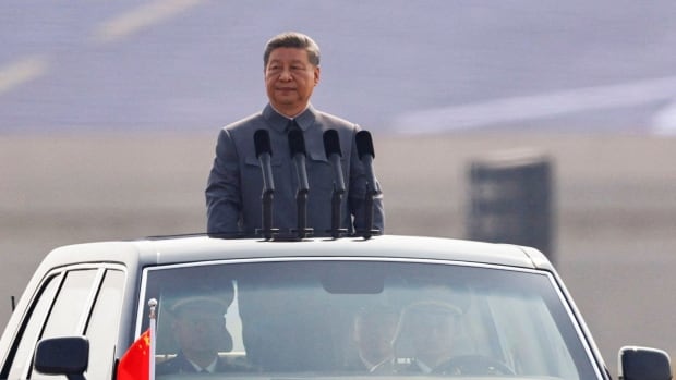 A man with black hair in a grey suit jacket stands with his head and shoulders out the top of a moving car