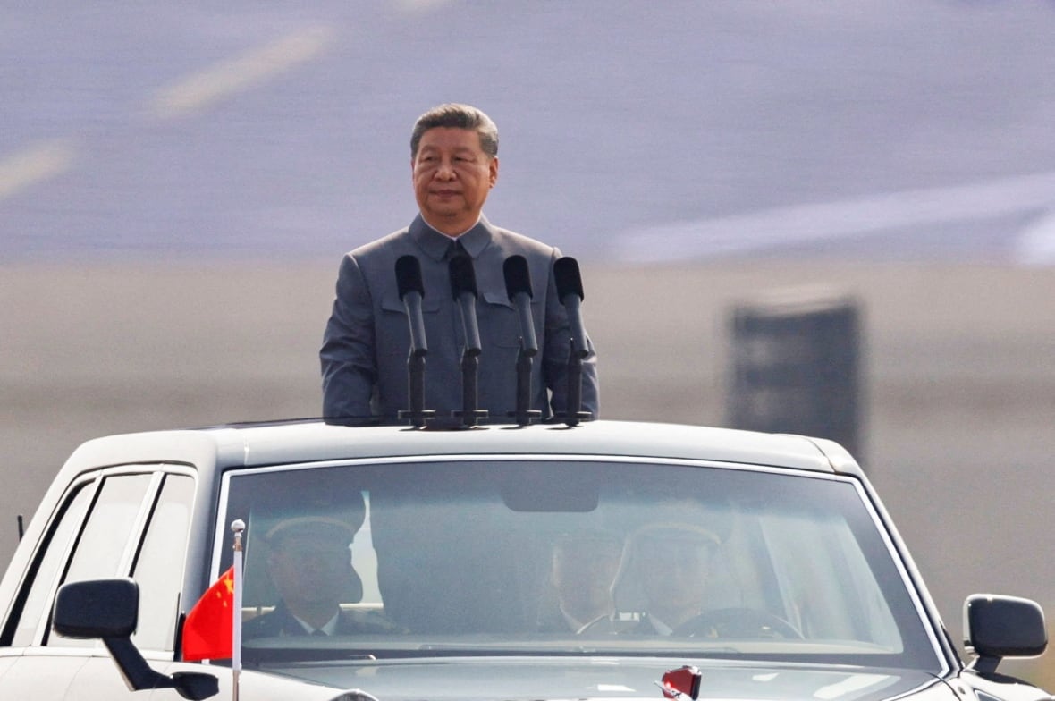 A man with black hair stands in a gray suit jacket and his head and leaves the top of a mobile car