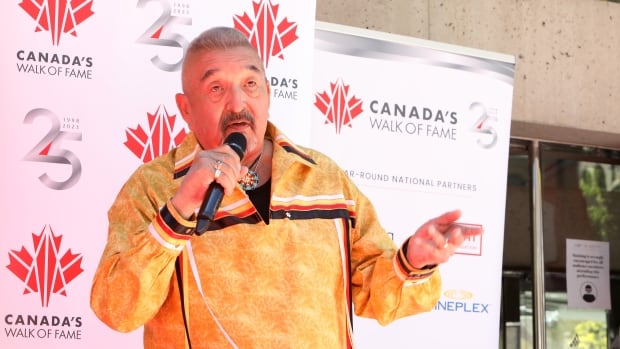 A man with short cropped grey hair and a mustache is shown speaking into a microphone while speaking at an outdoor event. 
