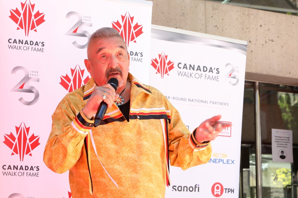 A man with short cropped grey hair and a mustache is shown speaking into a microphone while speaking at an outdoor event.