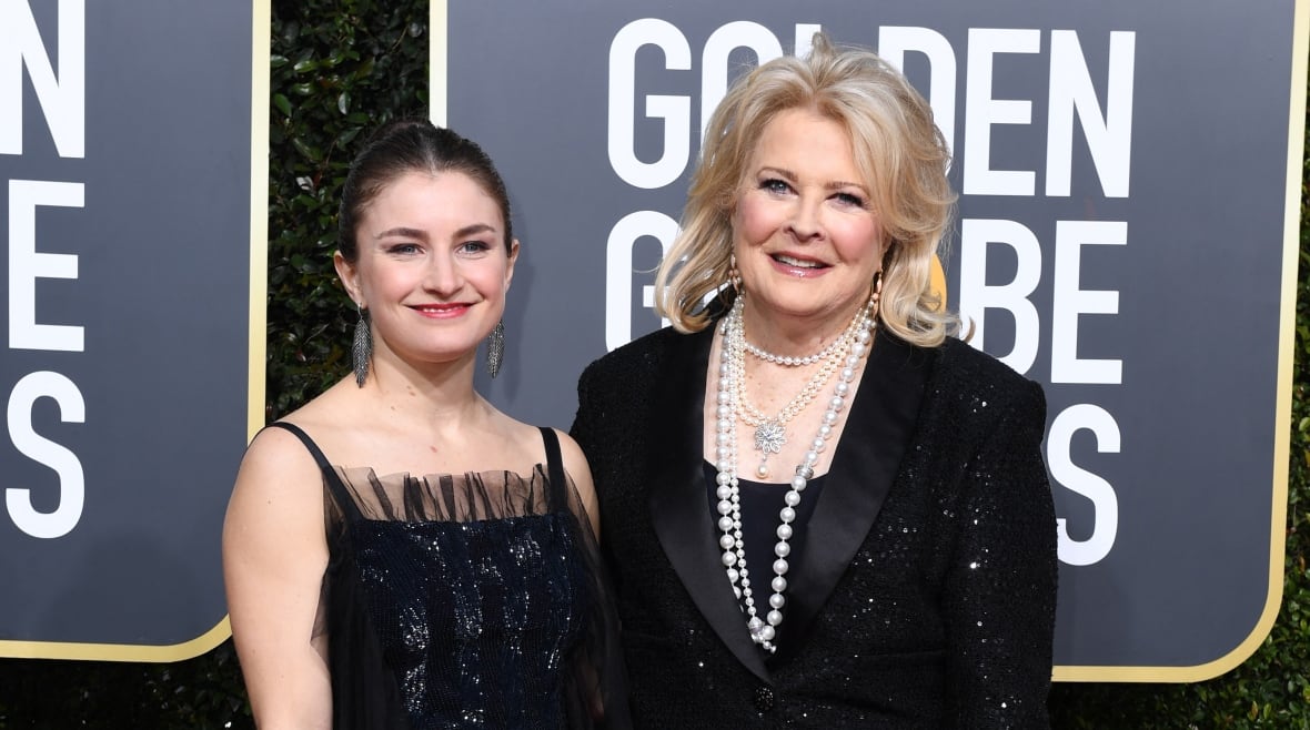 Two woman pose for a photo on a red carpet, one with dark hair pulled back in a bun and wearing a black sequin gown, the other with shoulder-length blonde hair and wearing a black sequin suit.