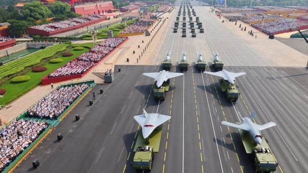 A bird's-eye view of a military parade as drones and other weapons pass spectators lining both sides of a multi-lane road.
