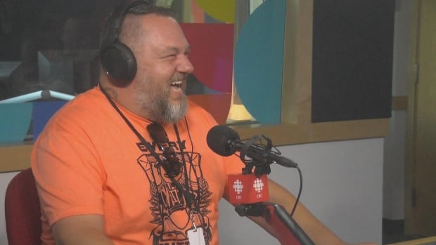 A man wearing an orange shirt laughs as he sits at a microphone
