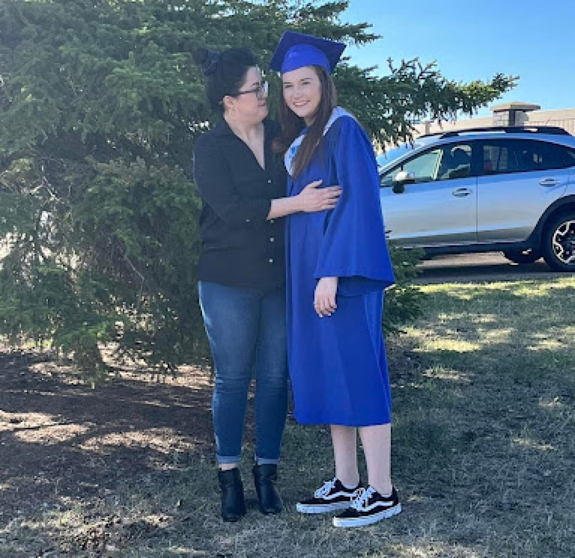 A young woman wearing a blue graduation cap and gown stands with another woman in front of a tree.