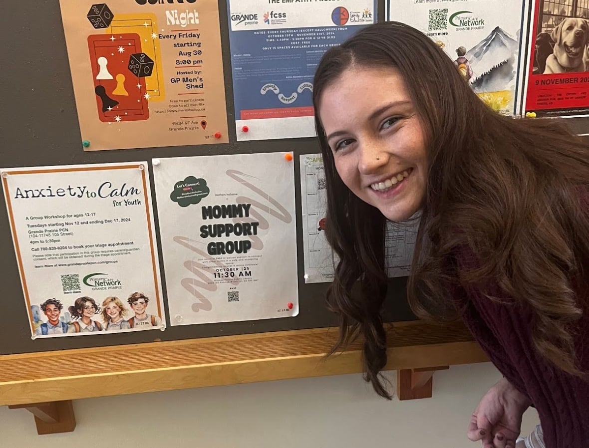 A smiling woman poses in front of a bulletin board. Beside her is a poster stating "Mommy Support Group"