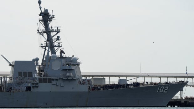 A grey warship with the number 102 on its hull sits in the water in front of a cloudy sky.