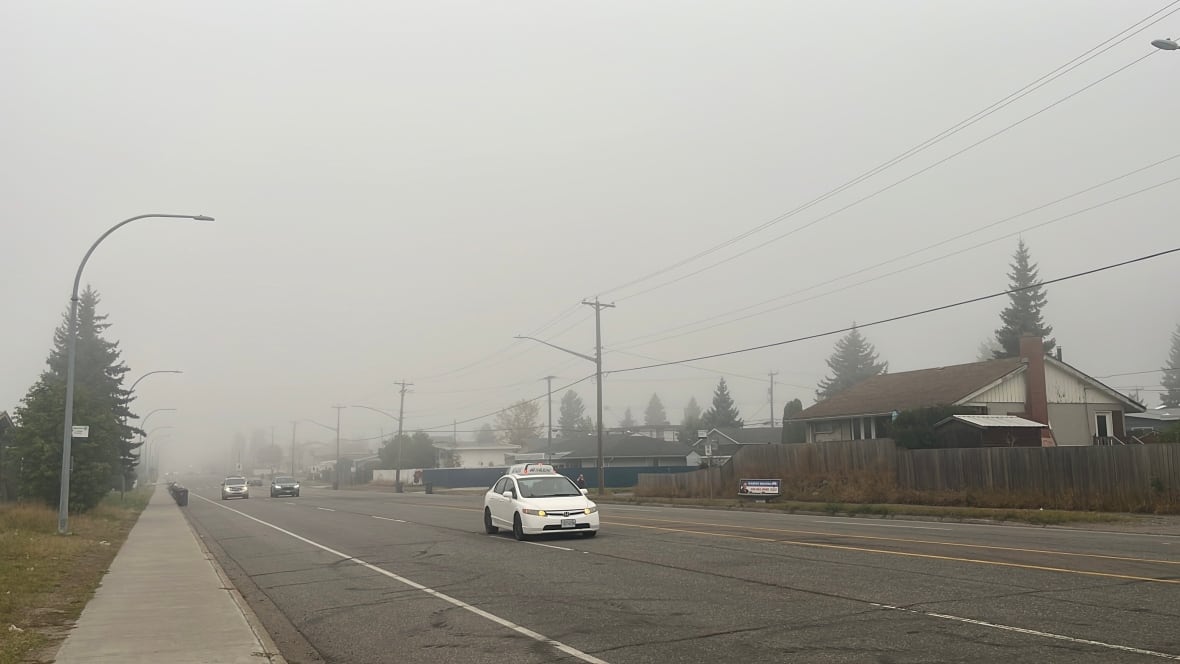 A white car drives down a four-lane road on a smoky day.