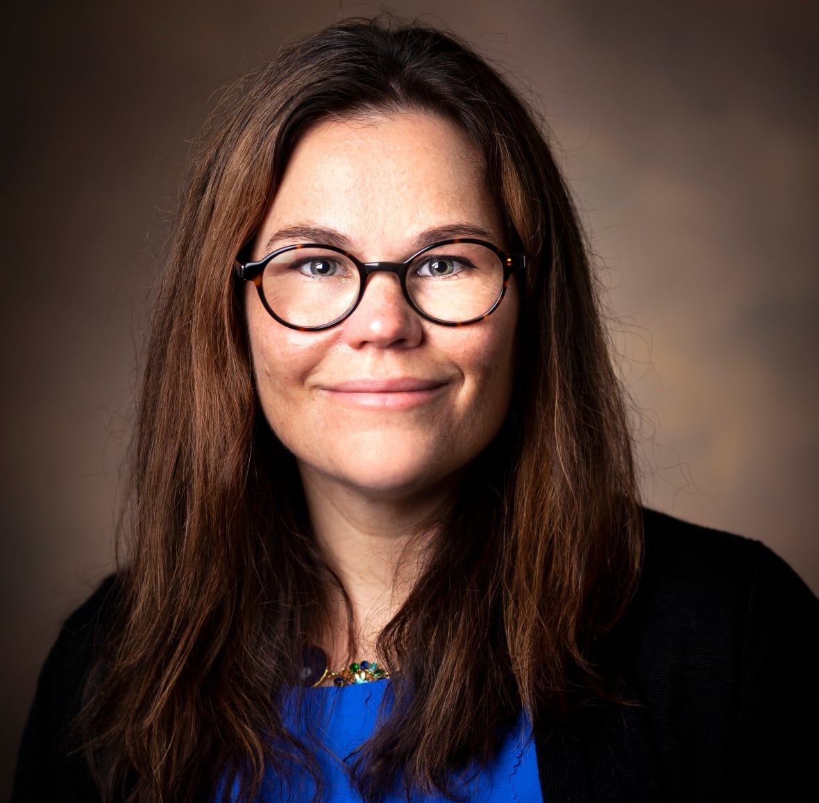 A woman with straight, brown hair past her shoulder smiles as she wears a dark blue blouse with a black jacket. 