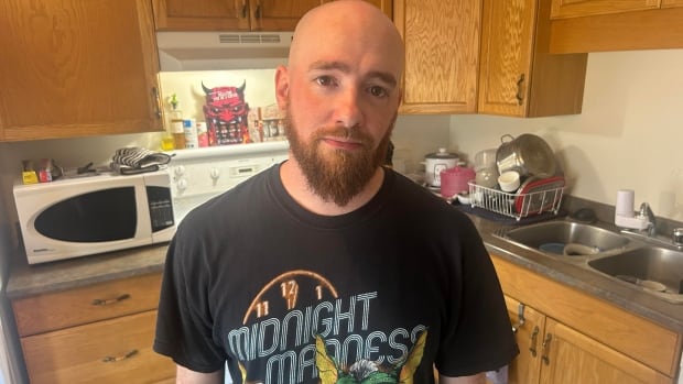 A bald man with a mustache and beard, wearing a black Midnight Madness T-shirt, stands in a kitchen.