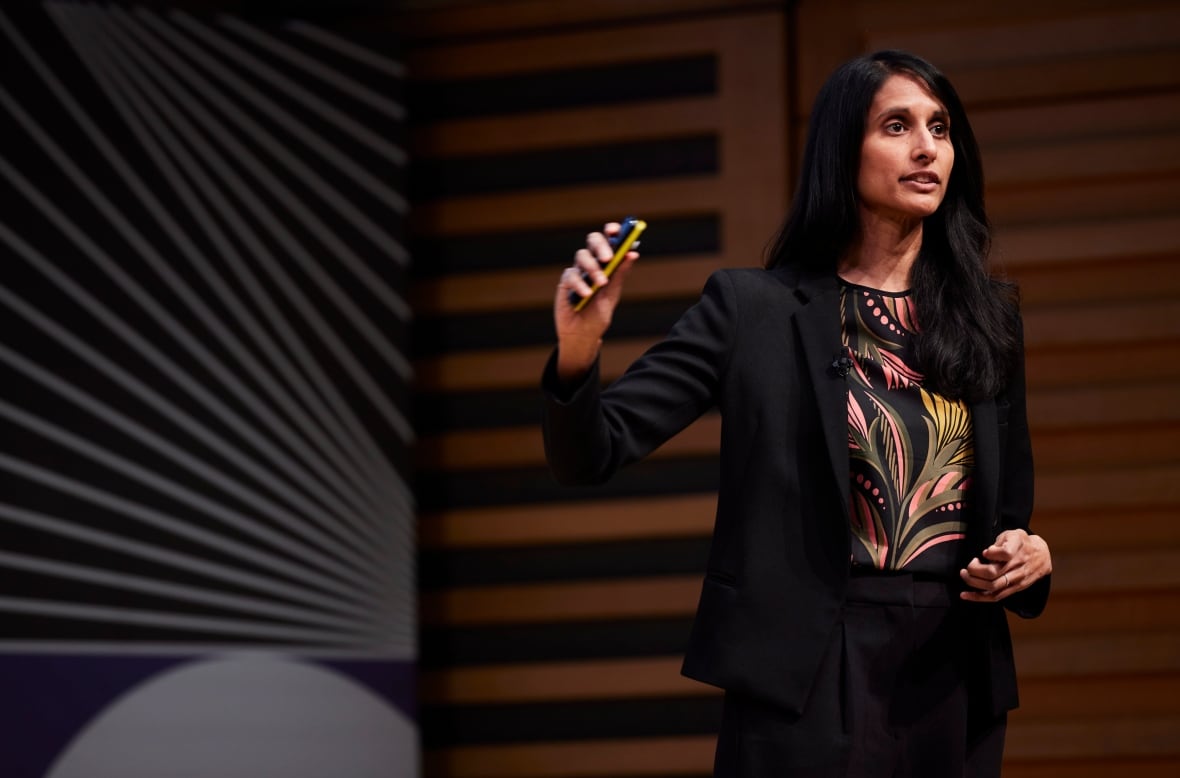 A woman standing with straight, black hair wearing a black suit, pink and yellow patterned shirt, uses a slide pointer.