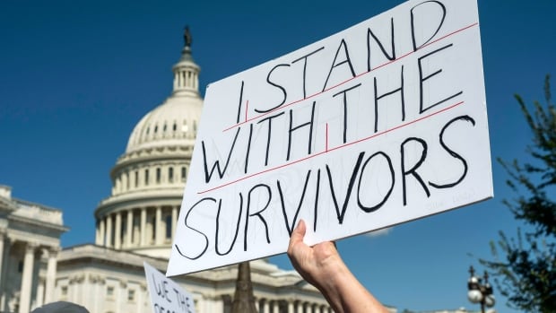 A white sign with black lettering reading I stand with the survivors, is held up against a backdrop of the Capitol Building
