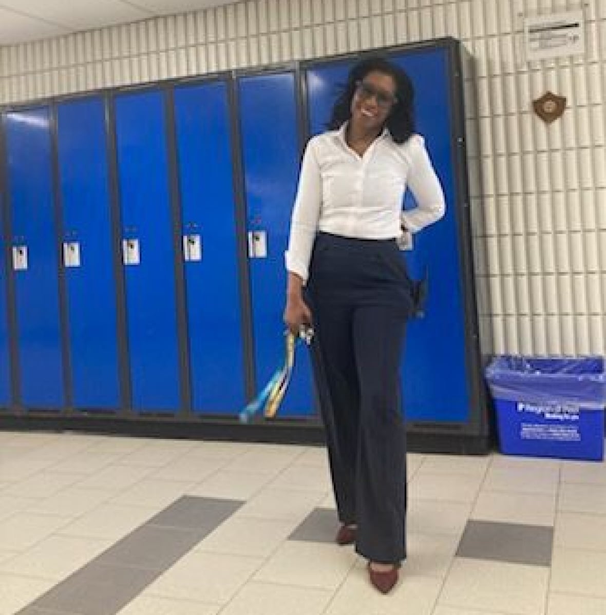 A woman wearing blue pants and a white shirt stands in a hallway with blue lockers.
