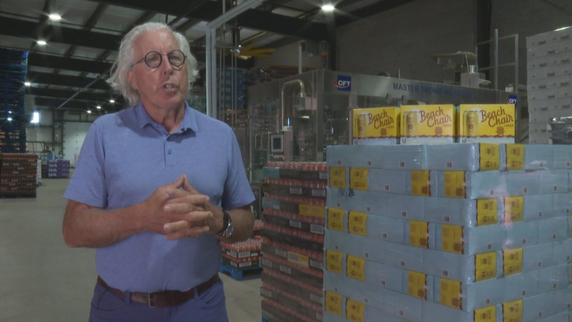 Man with white shoulder-length hair and glasses, wearing a blue shirt, stands in front of a pallet holding beer cans.
