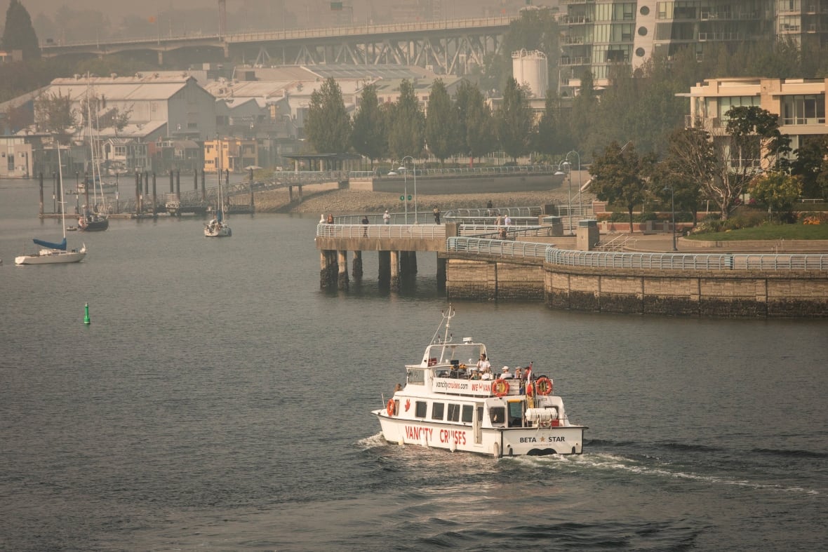 A pleasure boat passes by people on a nearby walkway in a water body, with industrial buildings visible in the background.