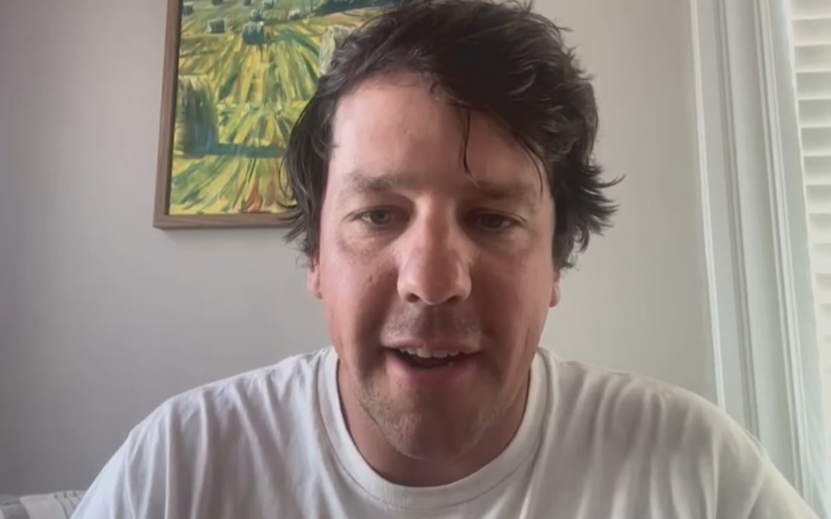 A man with dark hair in a white T-shirt Zooms from a room with a hayfield painting on the wall behind him.