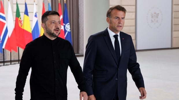 Two men, one cleanshaven and wearing a suit, and another bearded and in dark clothing, walk together in an indoor setting. 