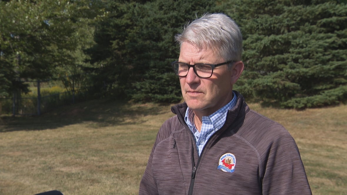 A man with gray hair and glasses wearing a brown fleece jacket with a small logo on it stands outside