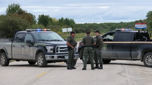 Three men wearing green uniforms stand on a road beside two black trucks. They are wearing vests that say "Conservation officer."