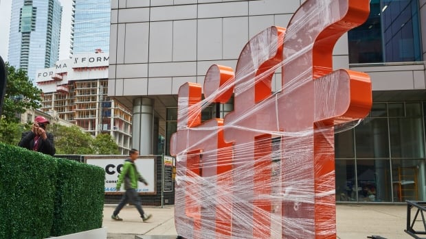 An sign consisting of four big, orange letters spelling out "tiff" sits wrapped in plastic on a city street as a pedestrian walks past in the background. 