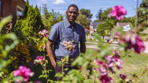 A Black man wearing a blue button up shirt stands behind a rose bush on a sunny day.