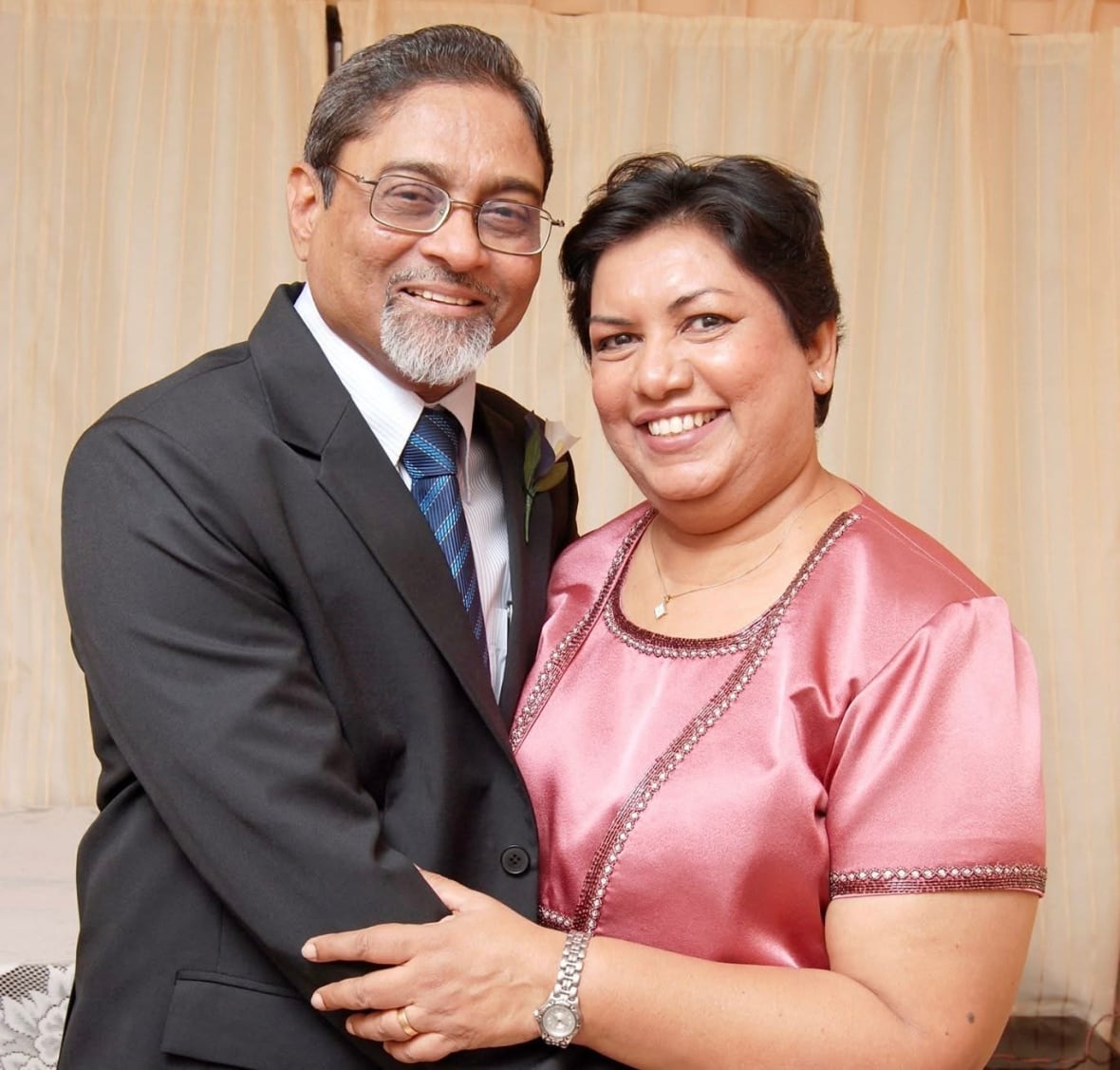 A middle -aged couple, the man with a suit and the woman who uses a pink satin outfit, hugs while smiling at a photo.
