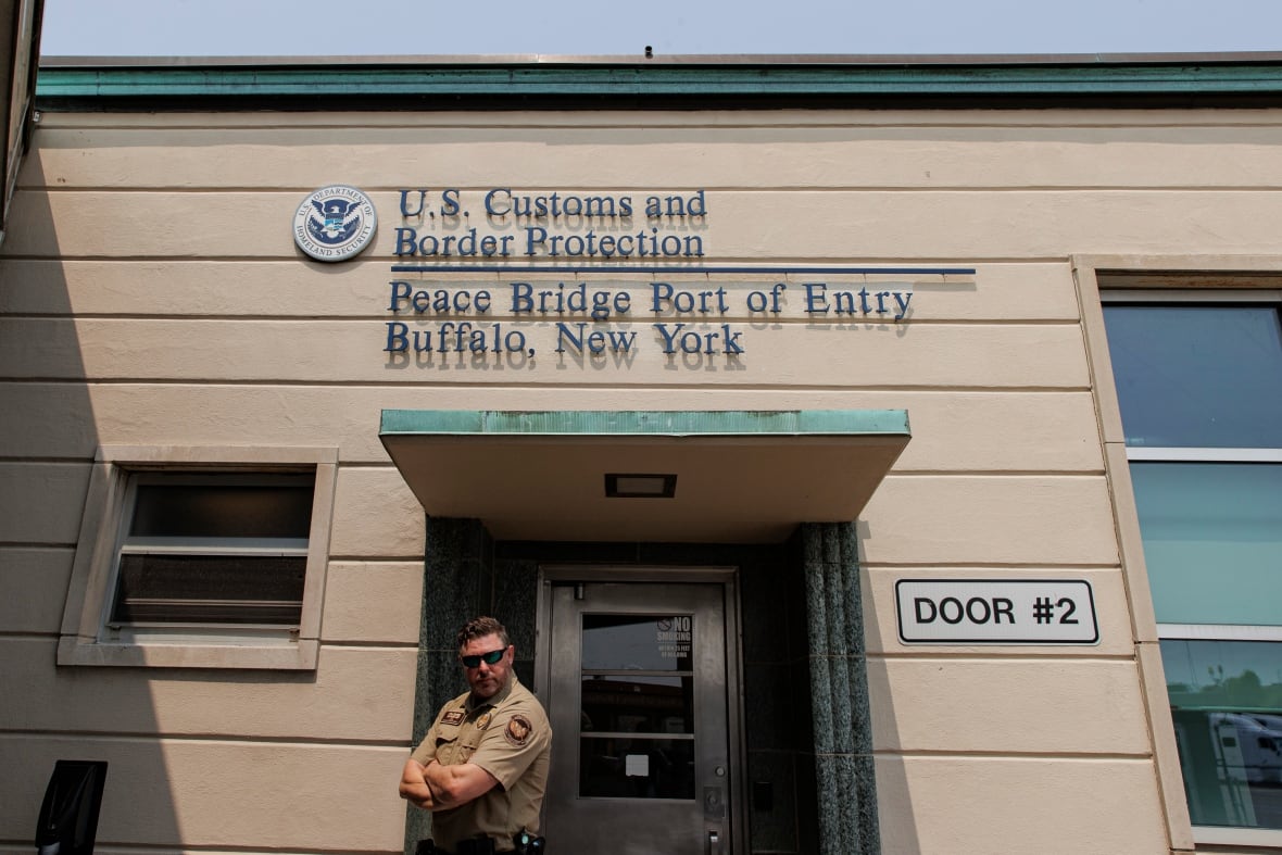 A man in a tan uniform and sunglasses stands in front of a tan building.