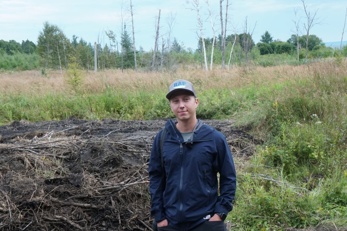 A man in a blue jacket and gray cap stands in front of a pile of mud and meadow