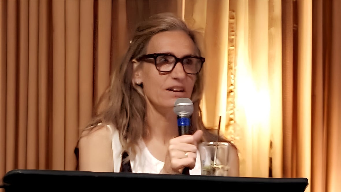 a women with blonde hair and glasses in a white shirt holds a microphone