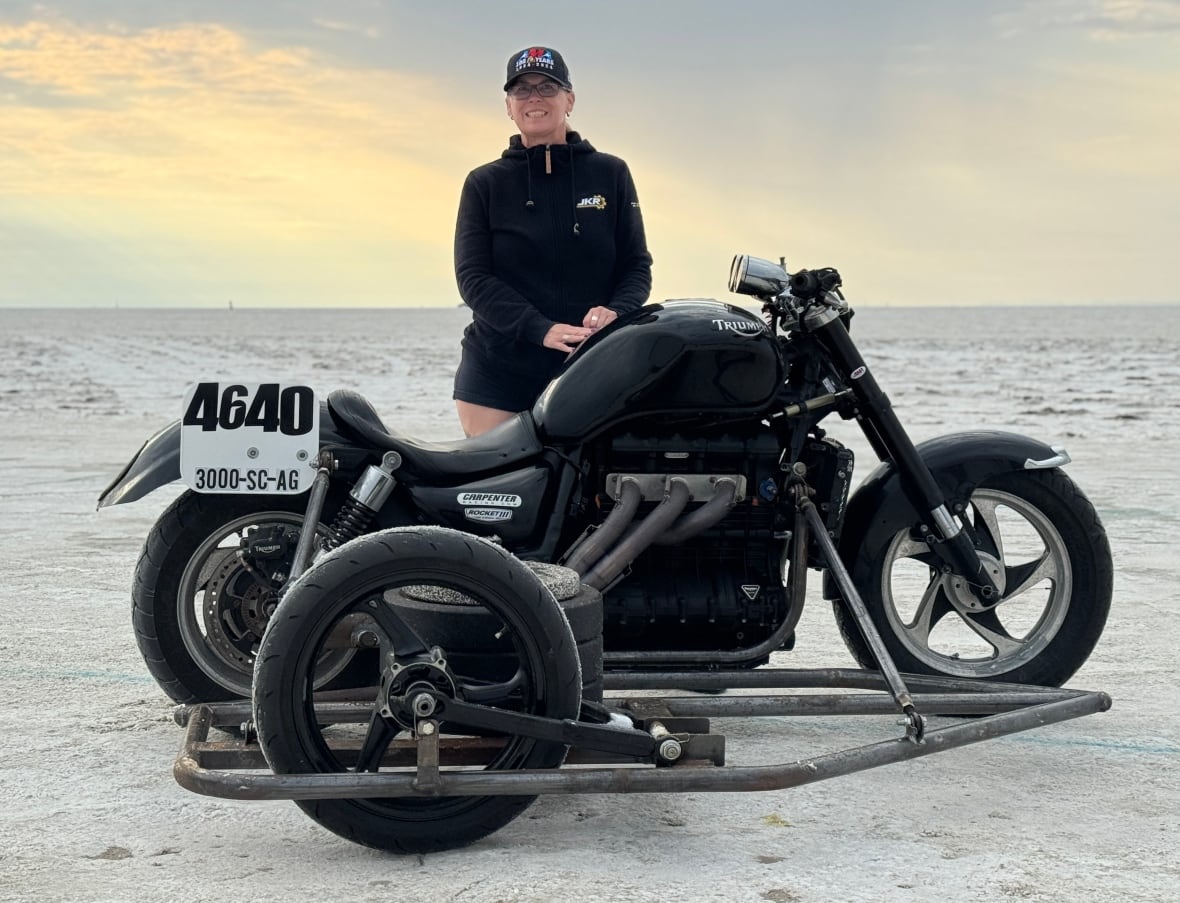 A woman in a black jacket and black baseball cap stands in front of a large black motorcycle with a sidecar attached to it.