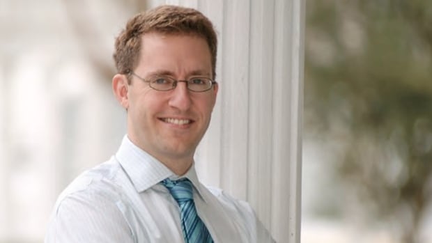 A man with short blond hair and glasses wearing a white dress shirt with a blue striped tie, crosses his arms over his chest and smiles for a portrait. 