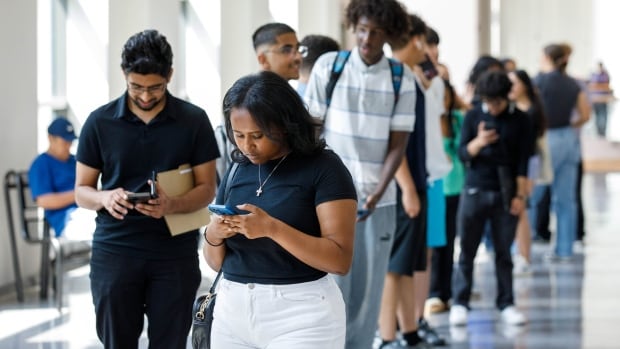 a woman in white shorts and a black tshirt looks down at her phone. shes the first person in a long line of young people standing behind her, waiting in a brightly lit hallways at a job fair