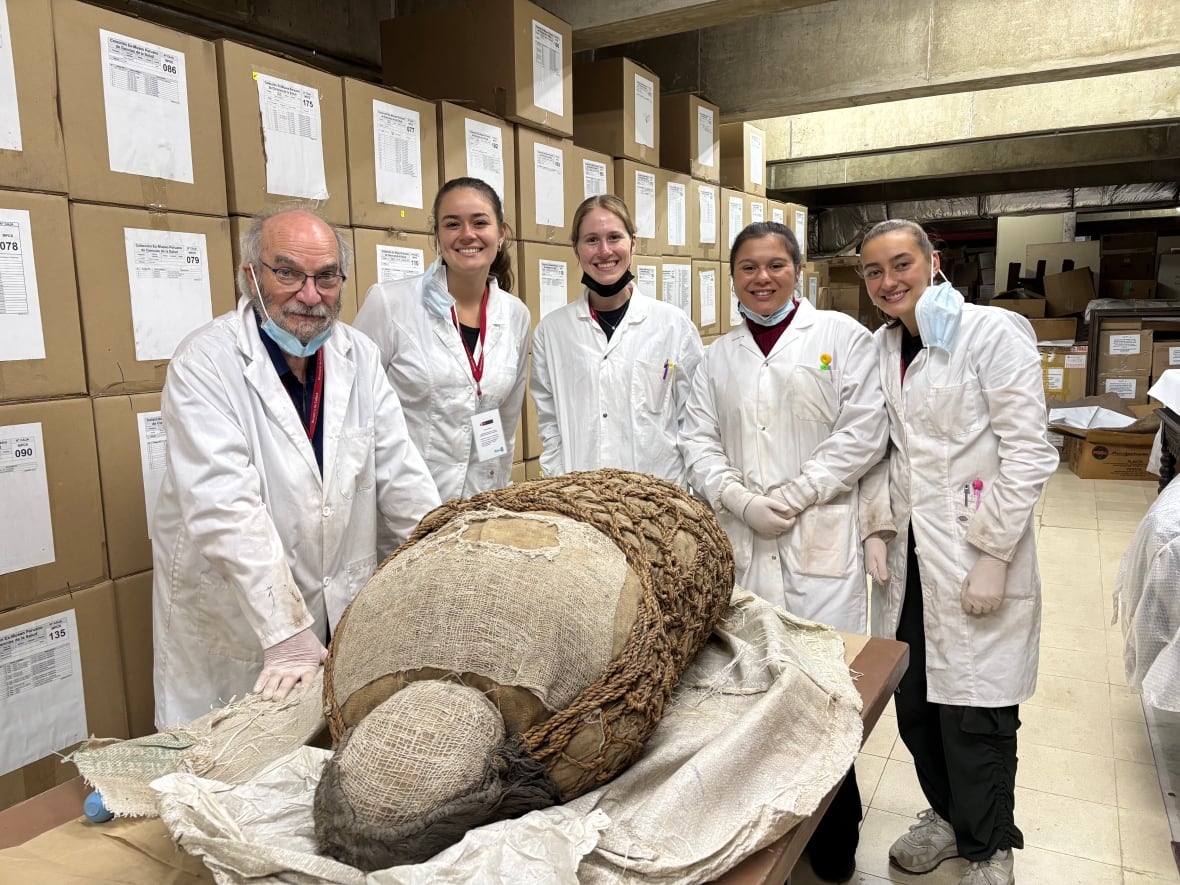 Five scientists, one older man and four younger females, stand in their lab coats around a table with a mummy on it wrapped in what looks like burlap.