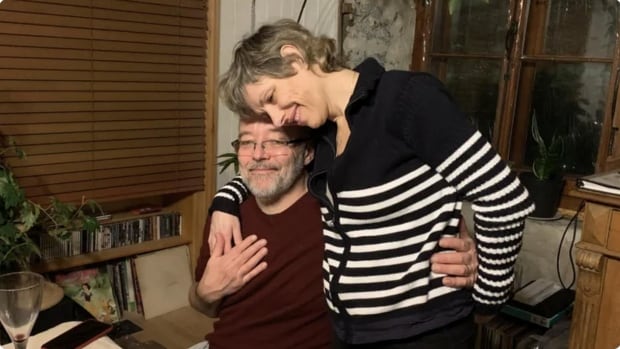 A pair of middle-aged adults embrace. A man is sitting while a woman leans over him. 