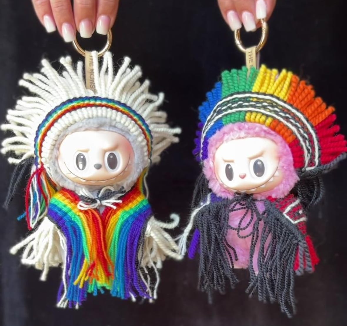 Well manicured hands hold up two little toy monsters by their keychain loops. Each is dressed in colourful hand-woven regalia, including headdresses.